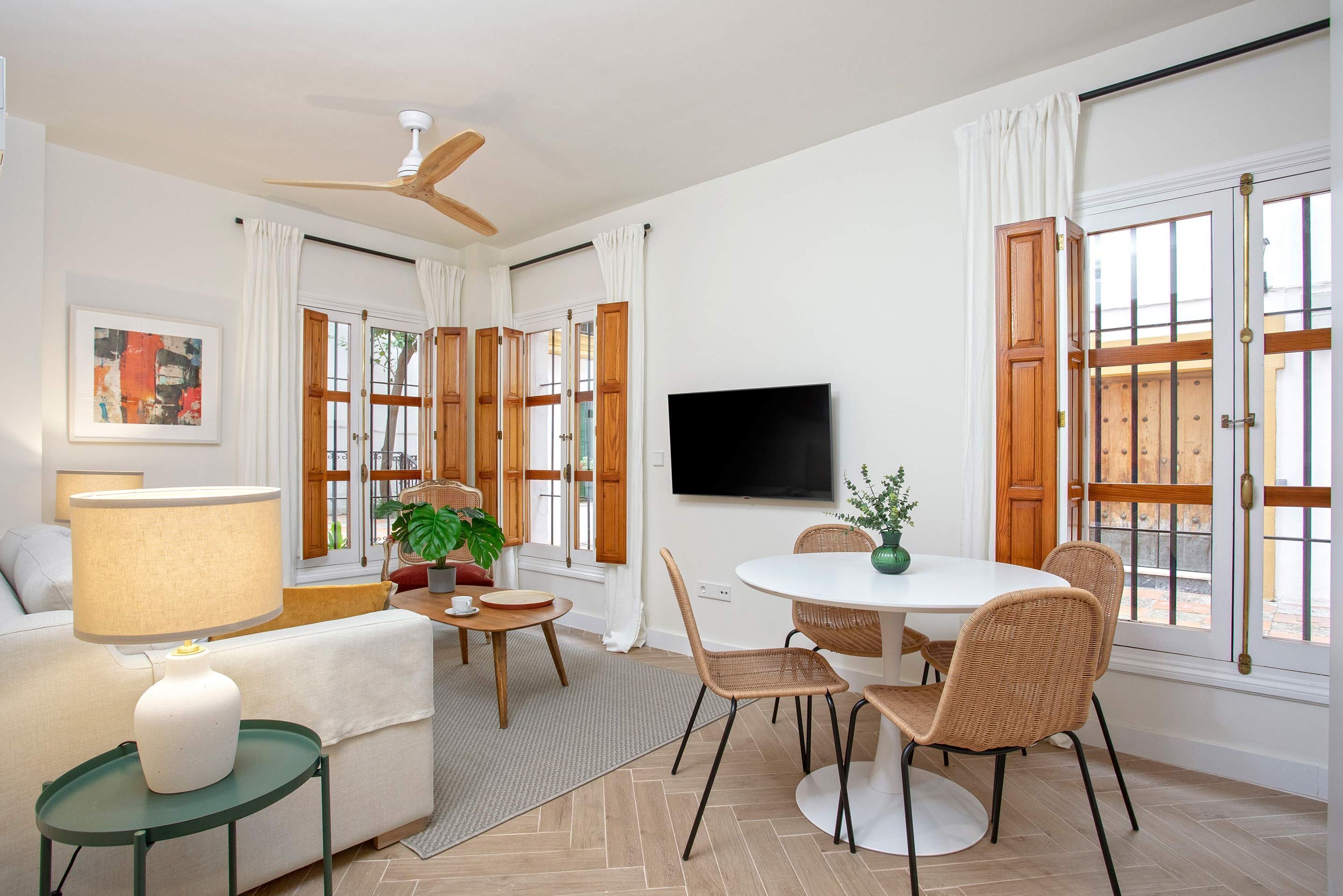 A modern living room with a sofa, a coffee table, a dining table with chairs, a TV mounted on the wall, and large windows with wooden shutters.