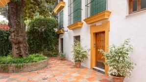 A courtyard with a checkered floor, potted plants, a tree, and a building with a wooden door and green shutters. - Bright Apartment in Seville Plaza Santa Cruz (Seville)