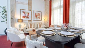 Un coin repas avec une table ronde dressée pour six personnes, entourée de chaises blanches. Des tableaux abstraux encadrés ornent le mur, une lampe se trouve sur une table d’appoint, et une grande fenêtre est agrémentée de rideaux rouges.