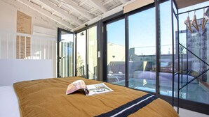Une chambre moderne avec un grand lit, un magazine posé sur le lit et une vue sur l’extérieur grâce à de grandes fenêtres.