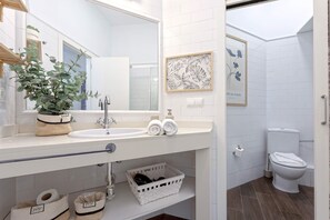 Ein modernes Badezimmer mit einem weißen Waschbecken, Spiegel und Toilette. Auf dem Waschbecken steht ein Topfpflanze, an der Wand hängt ein gerahmtes Blumenbild und darunter befindet sich ein weißer Korb.