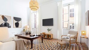 A modern living room with a geometric patterned floor, a wooden dining table, a sofa, a chair, and a television mounted on the wall. - Luxury Apartment in Malaga Larios VI (Málaga)