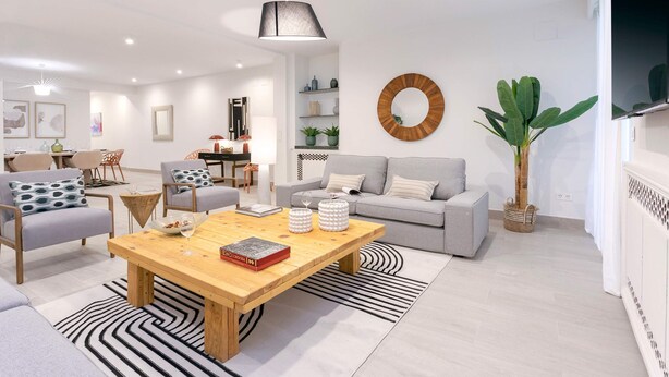 Ein modernes Wohnzimmer mit einem Holztisch, grauen Sofas und einer großen Pflanze.