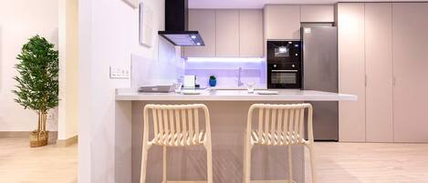 Uma cozinha moderna com uma ilha central, dois bancos altos, uma geladeira de aço inoxidável e um vaso com planta.