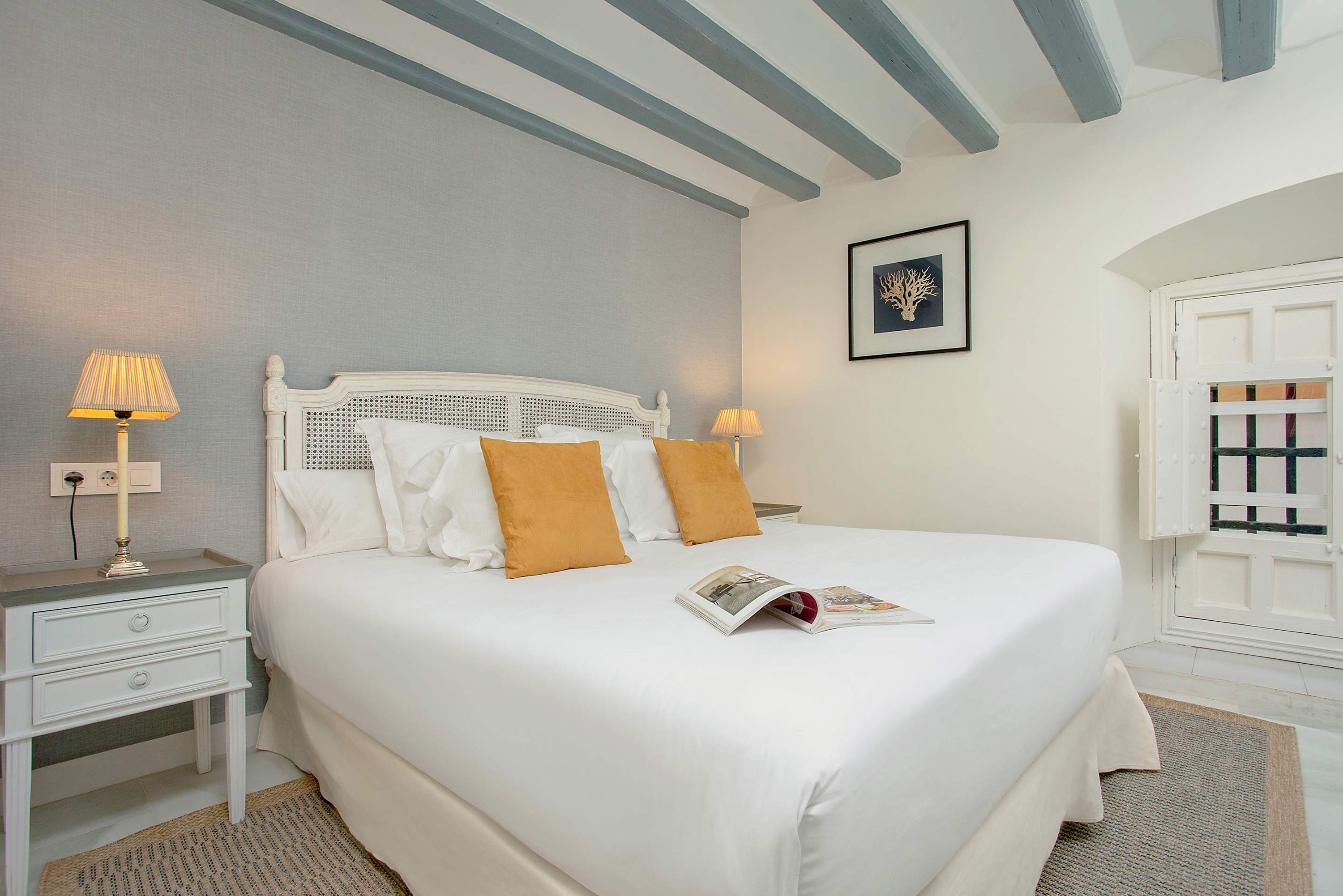 A neatly made bed with white linens and two orange pillows, a bedside table with a lamp, and a framed picture on the wall.