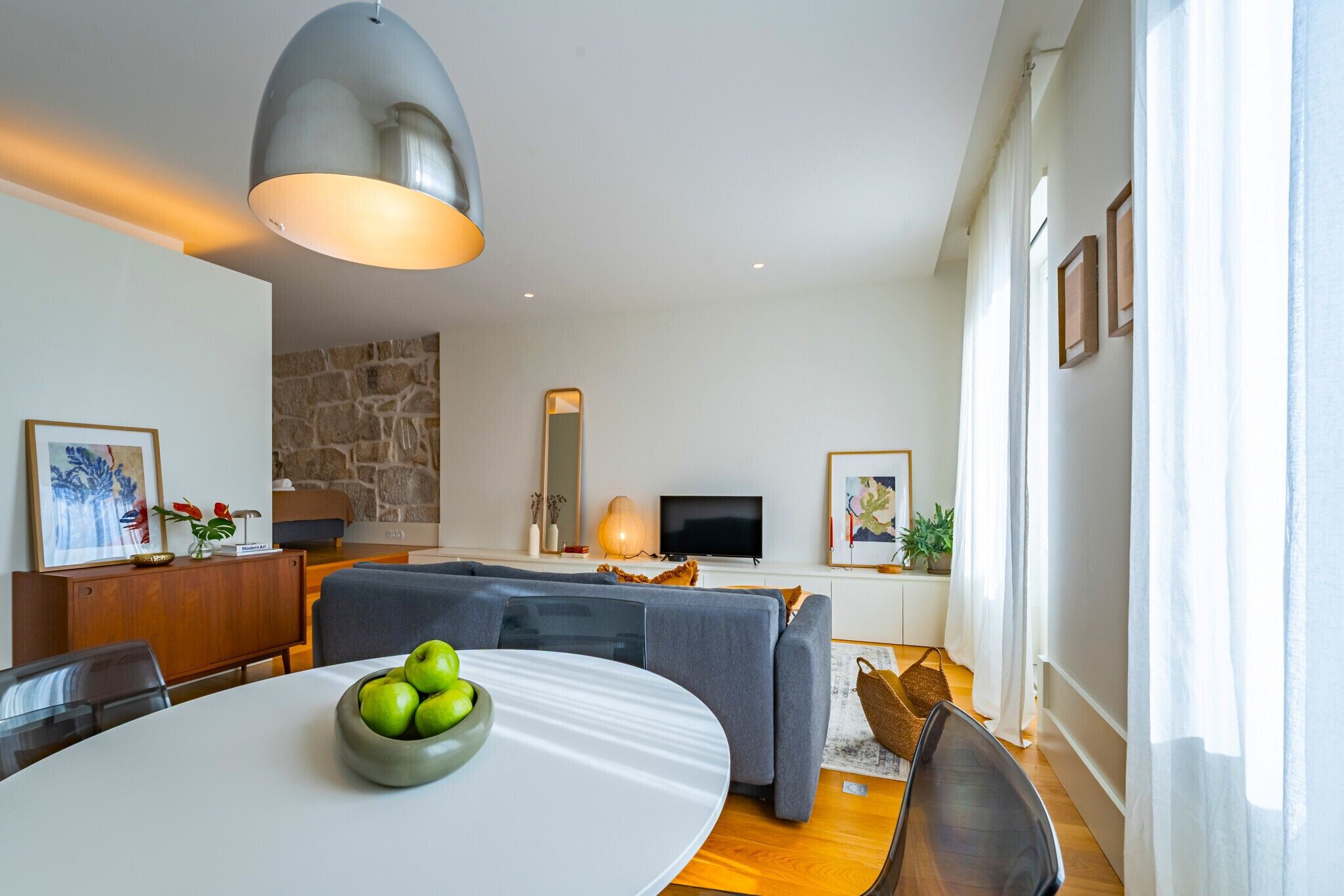 Ein modernes Wohnzimmer mit Esstisch, grauen Sofas, einer Wand mit Steinverkleidung, einem Flachbildfernseher und einem großen Fenster mit Vorhängen.