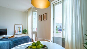 En moderne stue med en sofa, et spisebord med en skål æbler, et fjernsyn og et stort vindue med gardiner.