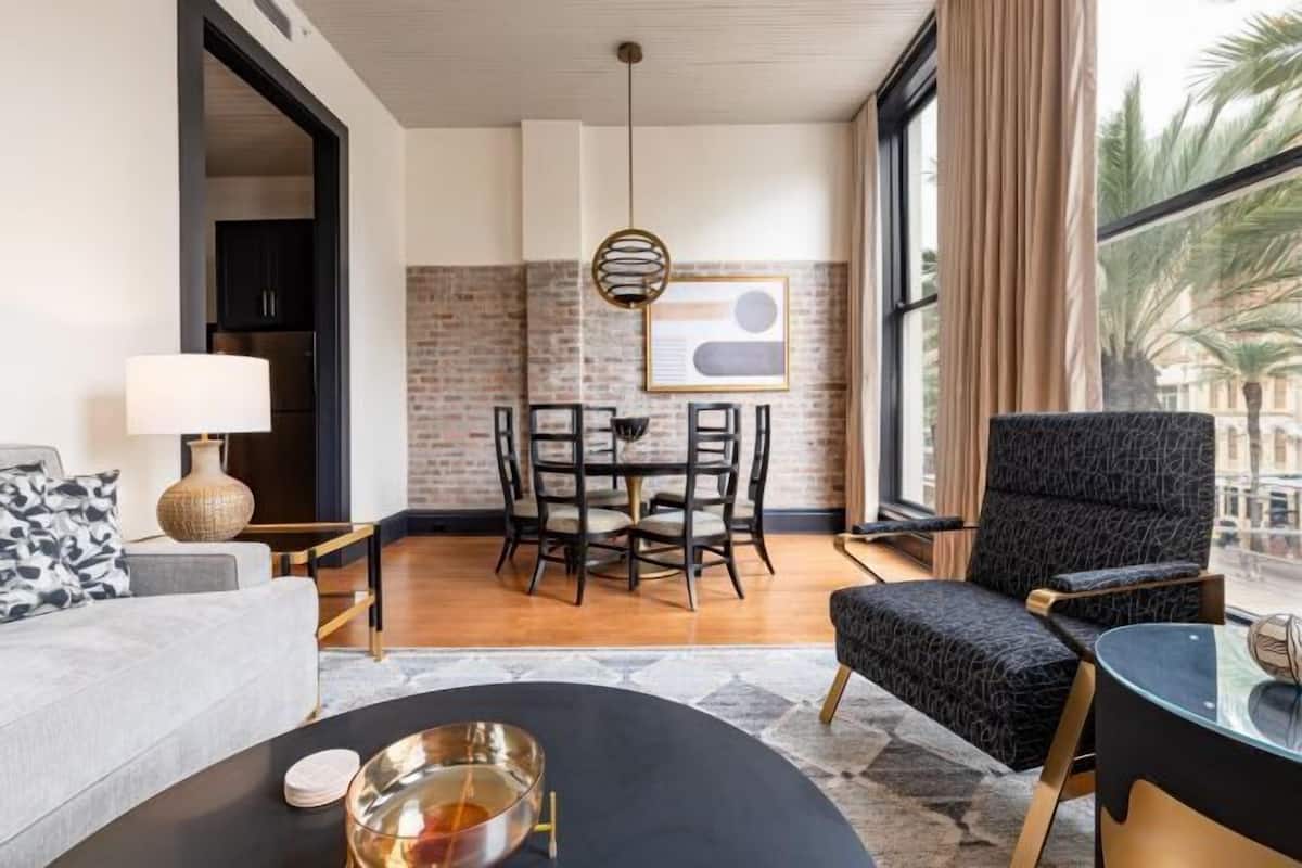 Un salon moderne comprenant un canapé, une table basse ronde, une table d’appoint avec une lampe, un coin repas avec une table et des chaises, et une grande fenêtre avec des rideaux.