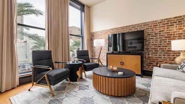 A modern hotel room with a large window, a brick accent wall, a flat-screen TV, and a round coffee table. - 2BR Luxury Stay & Stunning City Views The Mayfair (New Orleans)