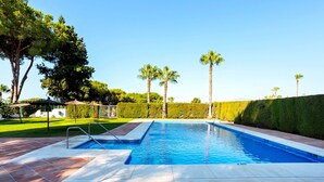 Una piscina rectangular con borde de baldosas, rodeada de palmeras y un seto.