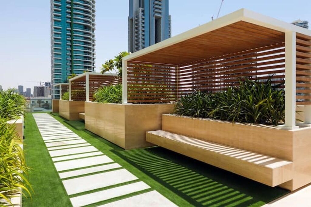 Un área de estar moderna al aire libre con estructuras de listones de madera y macetas, con edificios altos al fondo.