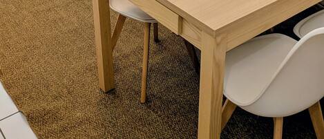 Una habitación con una mesa de comedor de madera, sillas blancas, un sofá beige con cojines verdes y un mantel individual tejido sobre la mesa.