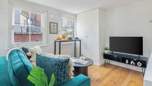 Una sala de estar moderna con un sofá color turquesa, un televisor de pantalla plana, una pequeña mesa redonda y un ventanal con vista a un edificio de ladrillo.