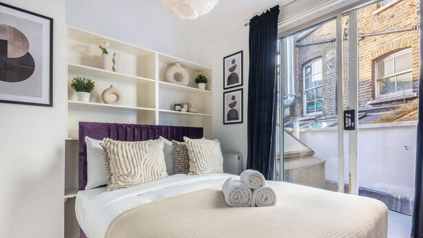 Una cama bien hecha con toallas enrolladas, un cabecero púrpura y una pared blanca con estantes que exhiben objetos decorativos.