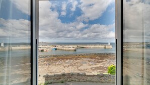 Blick durch ein Fenster auf die Küste, mit einem Sandstrand, einer Steinmauer und einem Steg mit Booten.