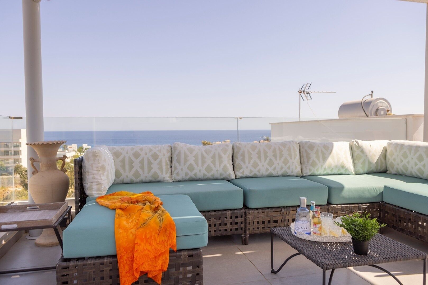 Eine Dachterrasse mit einem hellblauen Sofa, einem kleinen Tisch mit einer Pflanze und Blick aufs Meer.