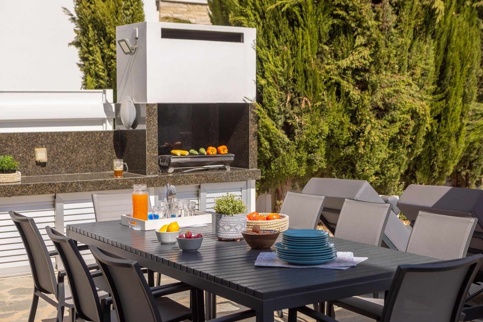 An outdoor dining area with a grill, table, chairs, and a basket of fruits.
