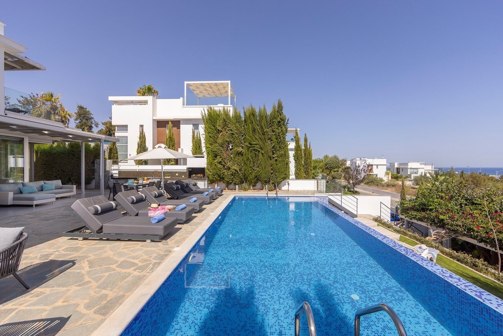 Un área de piscina moderna al aire libre con sillones de descanso y una parrilla, rodeada de árboles y edificios.