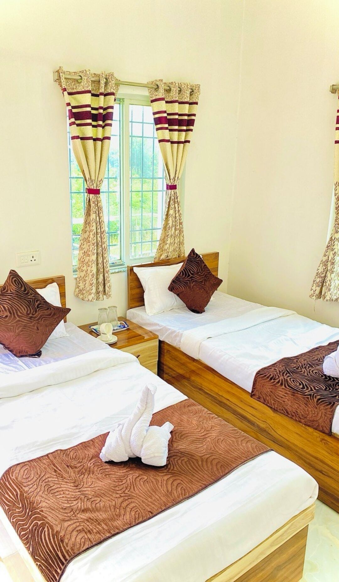 A hotel room with two single beds, a wooden headboard, a window with curtains, and a bedside table with a lamp.