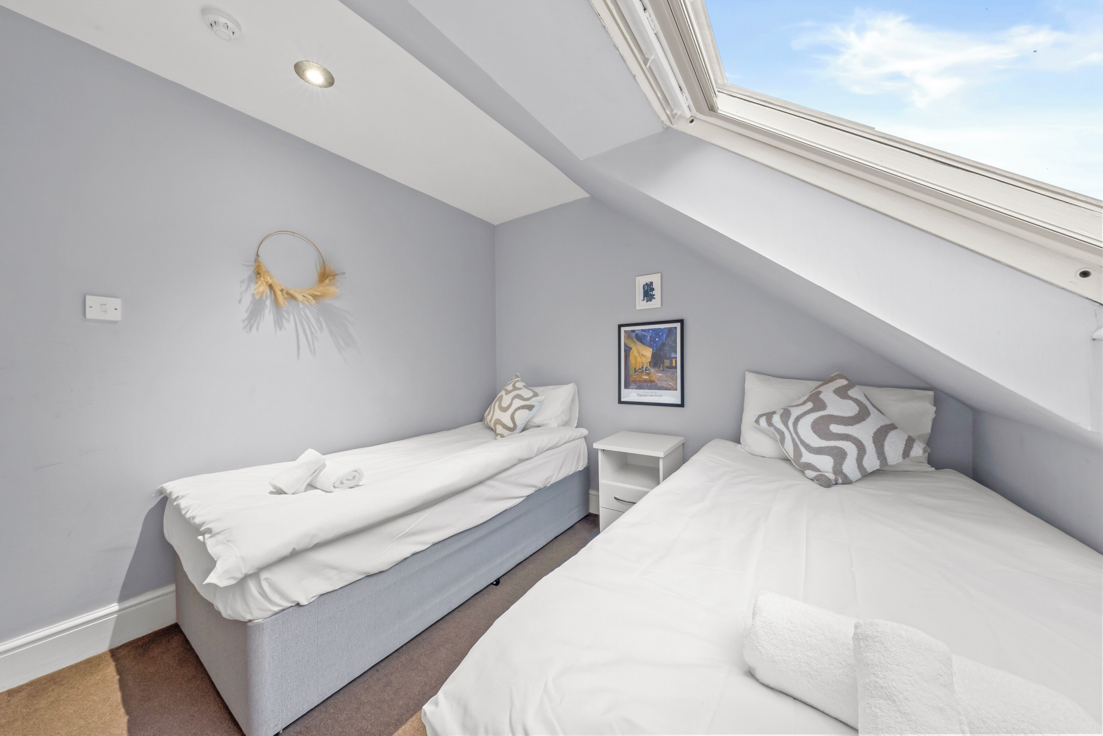A bedroom with two beds, a skylight, a wall-mounted artwork, and a small bedside table.