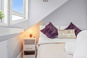 A bedroom with a white bed, two purple pillows, a beige pillow with "Snuggle" written on it, a bedside lamp, and a nightstand with a plant. - Charming Village Escape near Loch Lomond (Drymen)