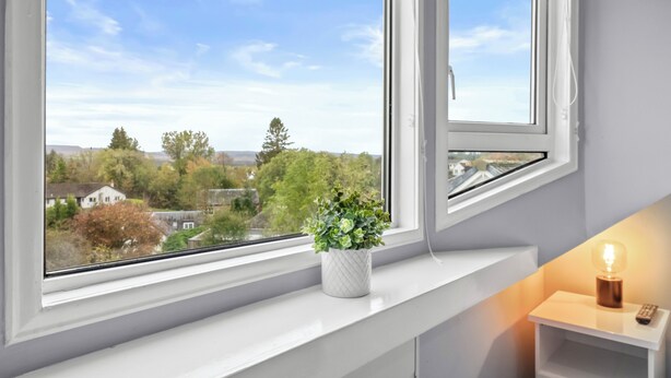 A room with a view of a cityscape through a window, a potted plant on the windowsill, and a bedside table with a lamp. - Charming Village Escape near Loch Lomond (Drymen)