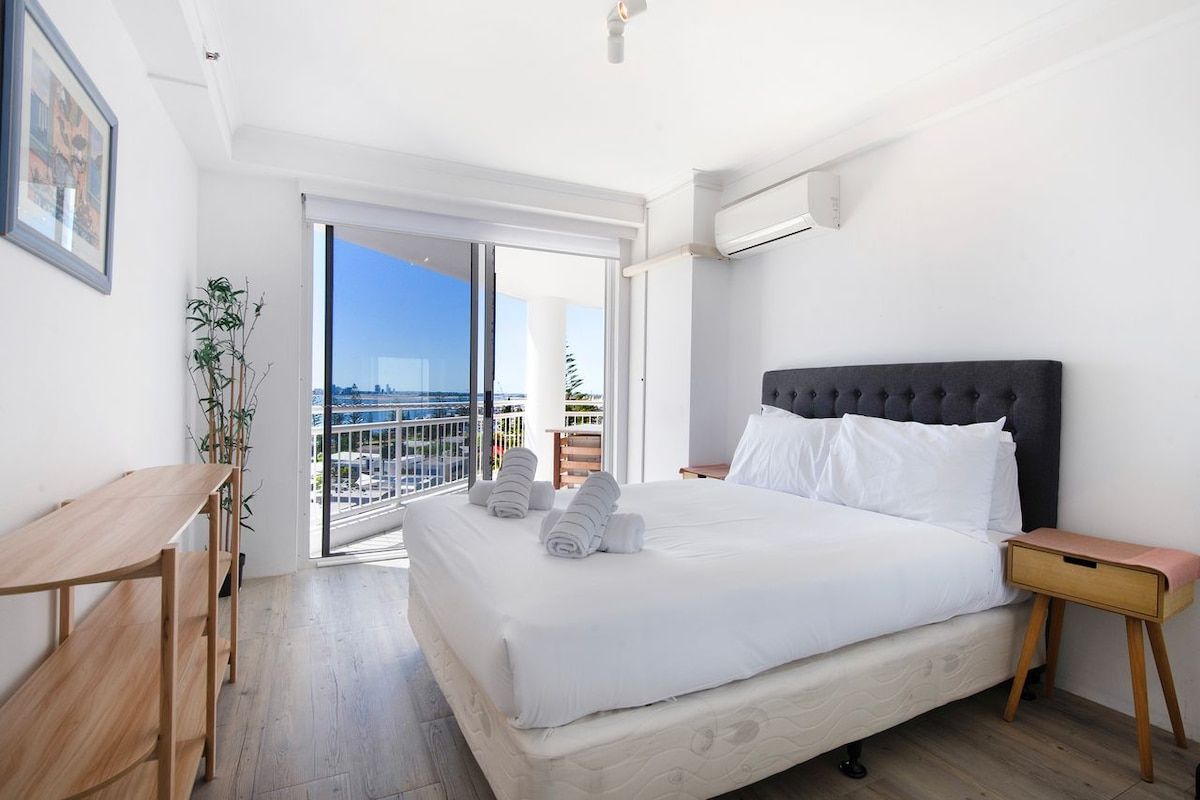 Habitación de hotel con cama, mesita de noche, un taburete pequeño, una planta en maceta y vistas a la ciudad a través de un ventanal grande.