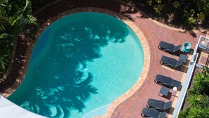 Eine Vogelperspektive auf einen Swimmingpool mit einer gefliesten Kante, umgeben von einer Pflasterterrasse und Liegestühlen.