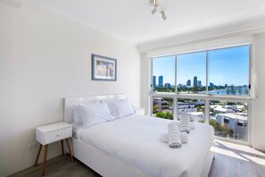 A modern bedroom with a large bed, a nightstand, a framed picture on the wall, and a city view through the window. - Ocean Sands Central Main Beach Apartment (Main Beach)