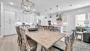 A modern kitchen with white cabinetry, a wooden dining table, and pendant lighting. - Beach Please by Stay on 30a (Inlet Beach)