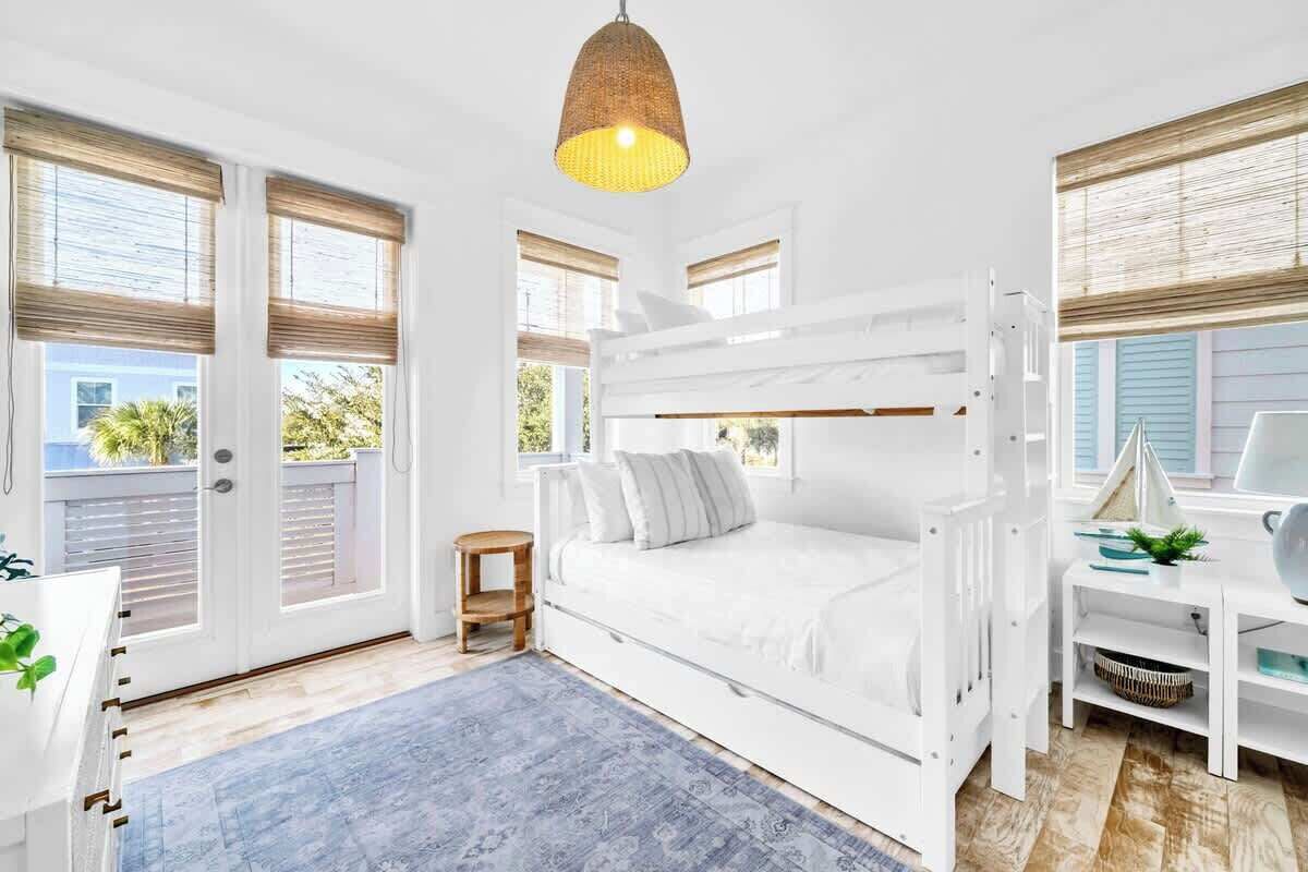 A white bunk bed room with a hanging light fixture, a small wooden stool, and a desk with a plant.