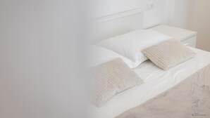 Ein Bett mit weißen Leinen und zwei beigen Kissen, ein weißer Nachttisch und eine schlichte Wand.