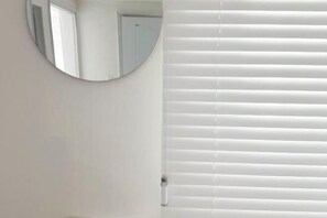 Un bagno con uno specchio rotondo, pareti bianche e una finestra con persiane orizzontali.