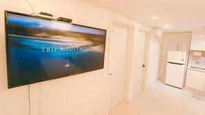 A modern hotel room with a large flat-screen TV mounted on the wall displaying 'TRIP MEDITATION PRESENTS'. The room has a minimalist design with white walls, a small kitchen area, and a door leading to another room. - Seoul Mapo 10people, 3 minutes DMC subway (Seoul)