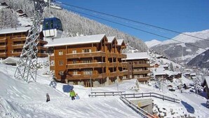 Ein Chalet-ähnliches Gebäude aus Holz mit mehreren Balkonen, einer Seilbahn im Hintergrund und schneebedeckten Bergen.