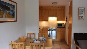 Eine kleine Küche mit Holzausstattung, einer Mikrowelle und einem Edelstahlbackofen. Ein Esstisch mit Stühlen und einer Schüssel sind ebenfalls vorhanden.