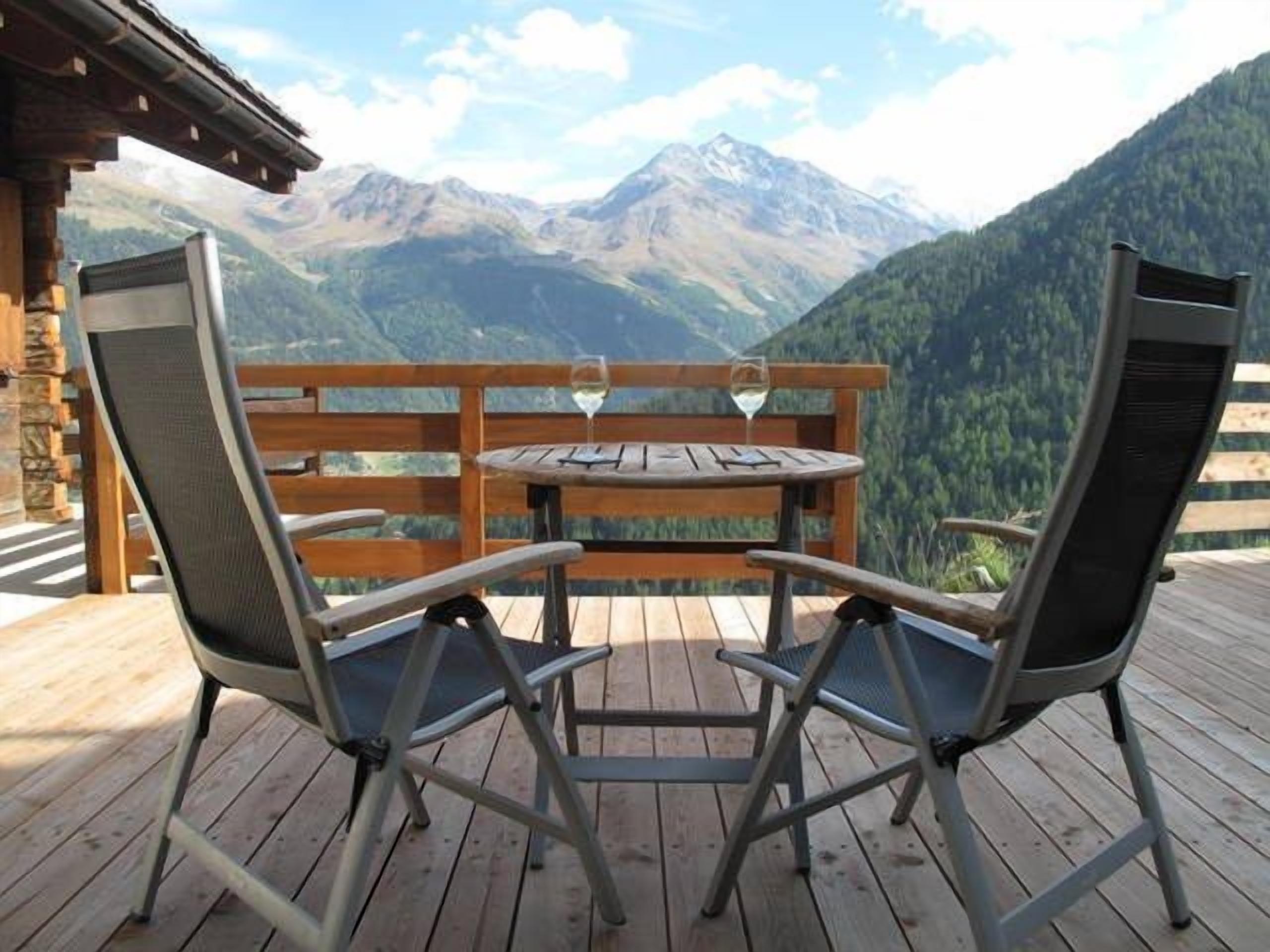 Um deck de madeira com duas cadeiras e uma mesa, com vista para uma paisagem montanhosa.