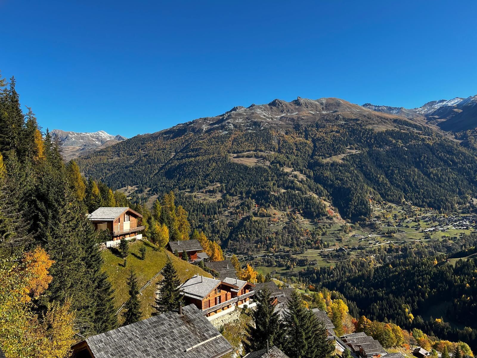 Un village de montagne aux maisons traditionnelles en bois, sous un ciel dégagé, et une vallée bordée de forêts denses et dominée par des sommets enneigés à l’horizon.