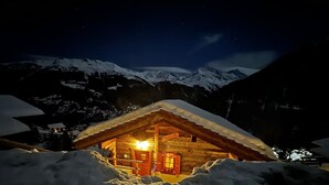 A cozy wooden cabin with a snow-covered roof, illuminated interior, and a view of snow-capped mountains under a starry night sky.