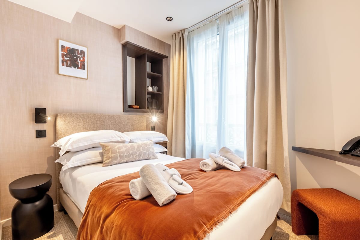 A hotel room with a bed, a bedside table, a nightstand, a built-in shelf, and a window with curtains.