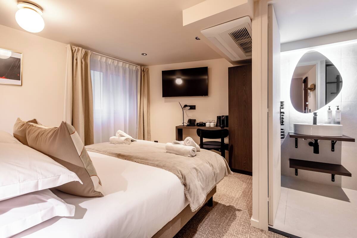 A hotel room with two beds, a TV, a desk, and a bathroom with a mirror and shelves.