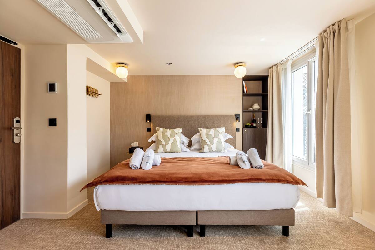 A modern hotel room with a large bed, a built-in heater, a bedside table, a nightstand, a bookshelf, and a large window with curtains.