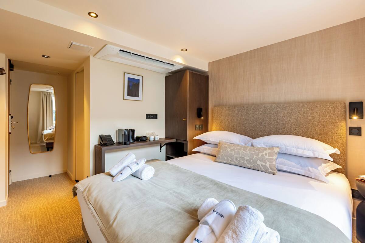 A hotel room with a large bed, a desk with a phone and a coffee maker, a mirror, and a wall-mounted air conditioner.