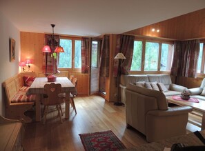 Ein gemütliches Wohnzimmer mit Holzboden, einer Couch, einem Esstisch mit Stühlen und einem Fenster mit Vorhängen.