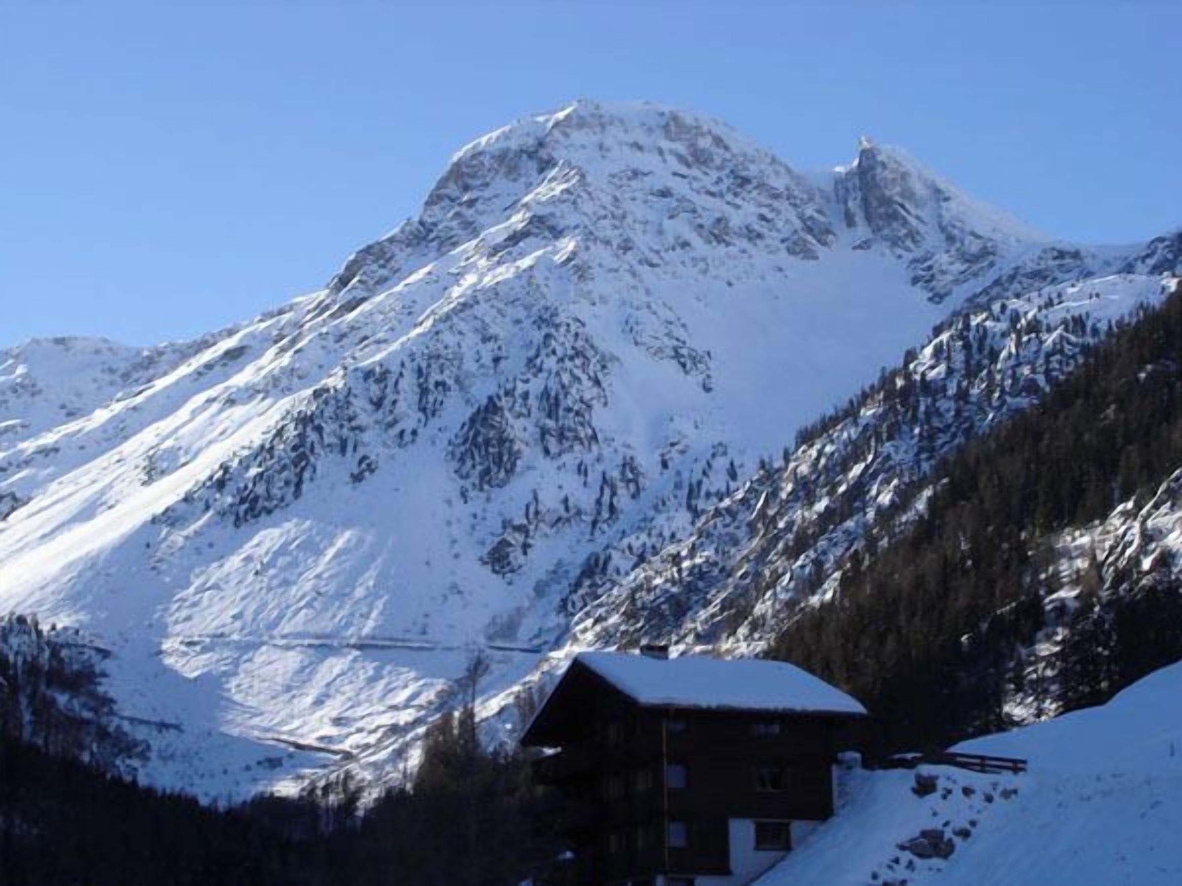 A mountain cabin with a snow-covered roof, surrounded by snow-covered slopes and a clear blue sky.