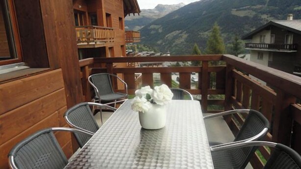 Un balcón de madera con una mesa y sillas, un florero con flores y vistas a la montaña.