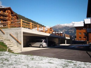 Un edificio con una escalera de madera que da a un estacionamiento techado, un automóvil estacionado en su interior y una montaña al fondo.