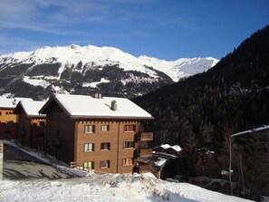A multi-story building with a snow-covered roof, set against a backdrop of snow-capped mountains and dense forest. - Very comfortable 4-room apartment for 6 persons : Living room - kitchen with dishwasher, Microwave a (Grimentz)