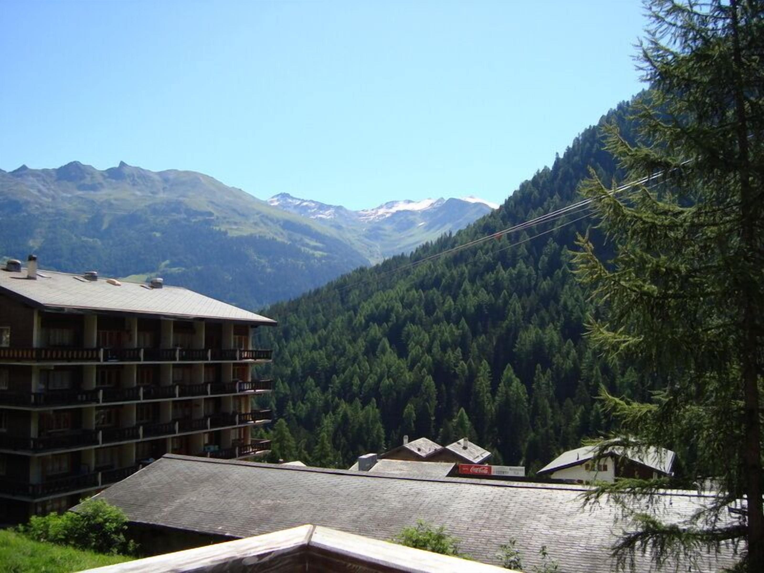 A multi-story building with balconies, surrounded by dense forest and mountains in the background.