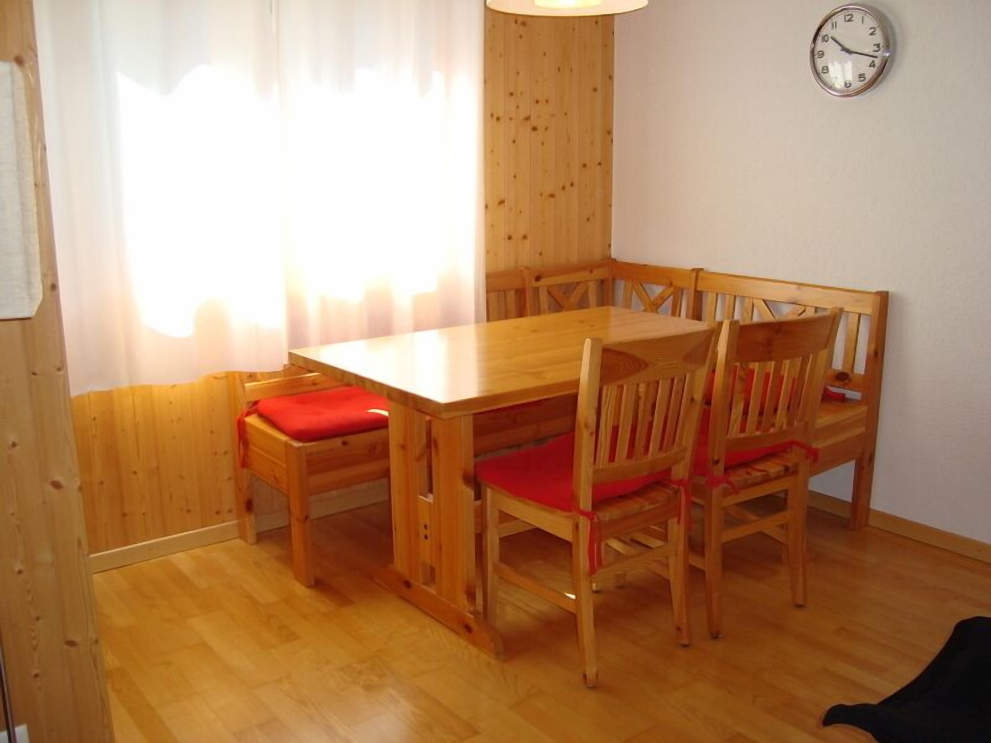 A wooden dining table with six chairs, a clock on the wall, and a window with curtains.